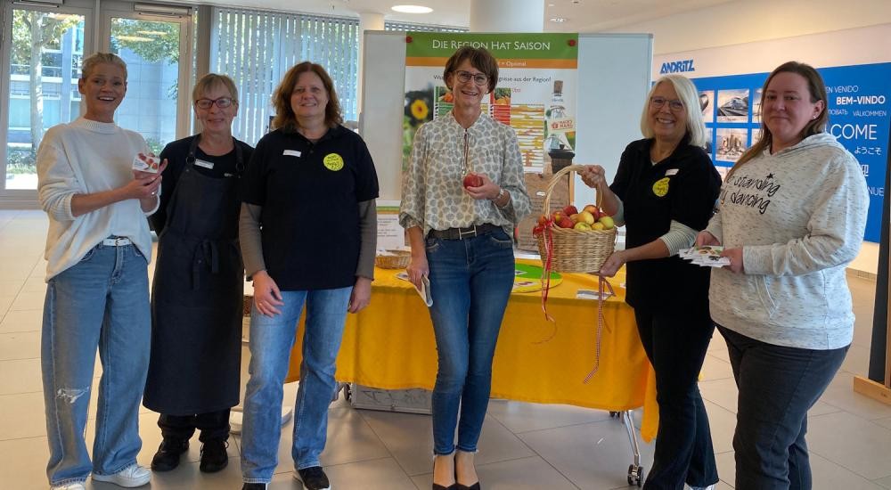 Vier Mitarbeiterinnen des Unternehmens ANDRITZ Schuler und zwei Kolleginnen aus der Abteilung Hauswirtschaft und Ernährung des Landwirtschaftsamtes am Informationsstand die sich einen Apfel aus einem Korb aussuchen .