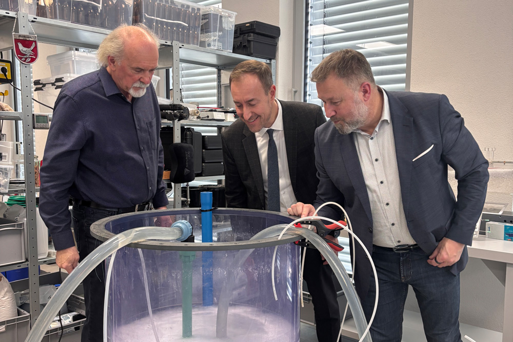 Beusch im Labor der Firma IEG. Drei Personen stehen zusammen an einer Maschine in Form eines großen Zylinders mit Schläuchen und Wasser darin (Umwelttechnik).