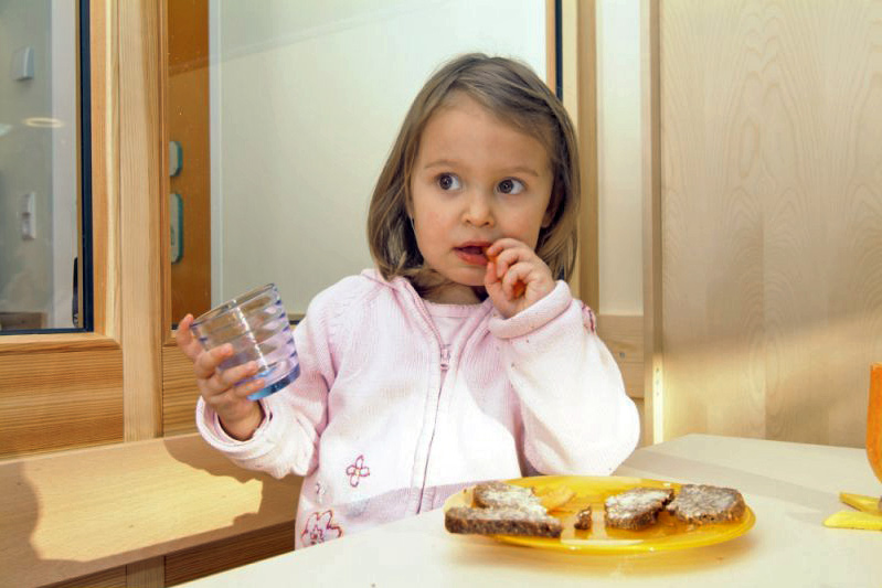 Kleinkindernährung Kind in rosa Jacke mit Glas in der Hand, auf dem Tisch ein Teller mit Brot