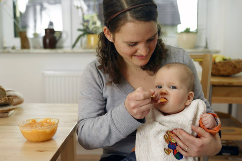 Start in die Breikost Mutter mit Säugling auf dem Schoß füttert Brei
