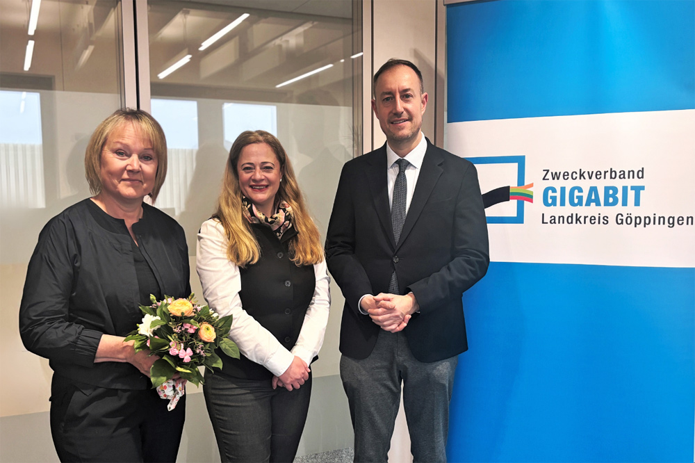 Drei Personen stehen vor einem Banner des Zweckverbands Gigabit. Links steht die neue Geschäftsführerin mit einem Blumenstrauß in der Hand.