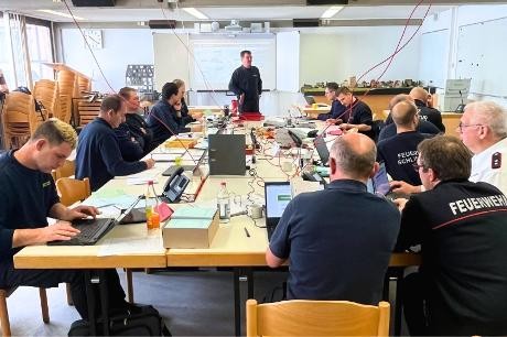 Übung des Feuerwehr-Führungsstabes im Landkreis Göppingen Mehrere Feuerwehrleute sitzen an einem langen Tisch mit Laptops, Telefonen und Kabeln, während eine Person vorne vor einem Bildschirm steht und spricht.