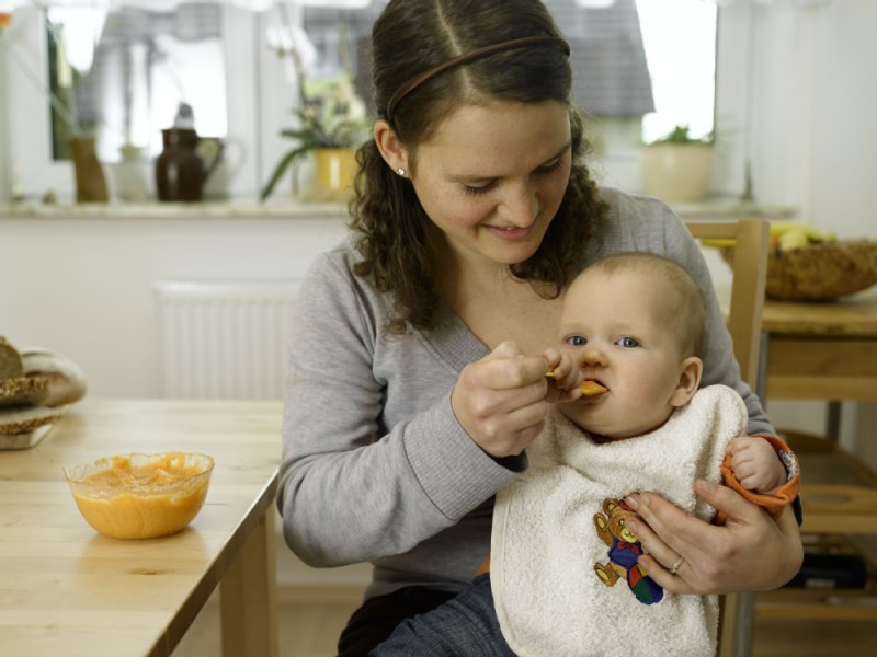 Der erste Brei Baby sitzt auf dem Schoss, Mutter füttert es mit Löffel
